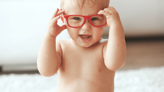 A baby wearing red glasses is standing on a white carpet.